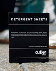 Black detergent sheet box with white writing in front of background of a city sidewalk and people walking.
