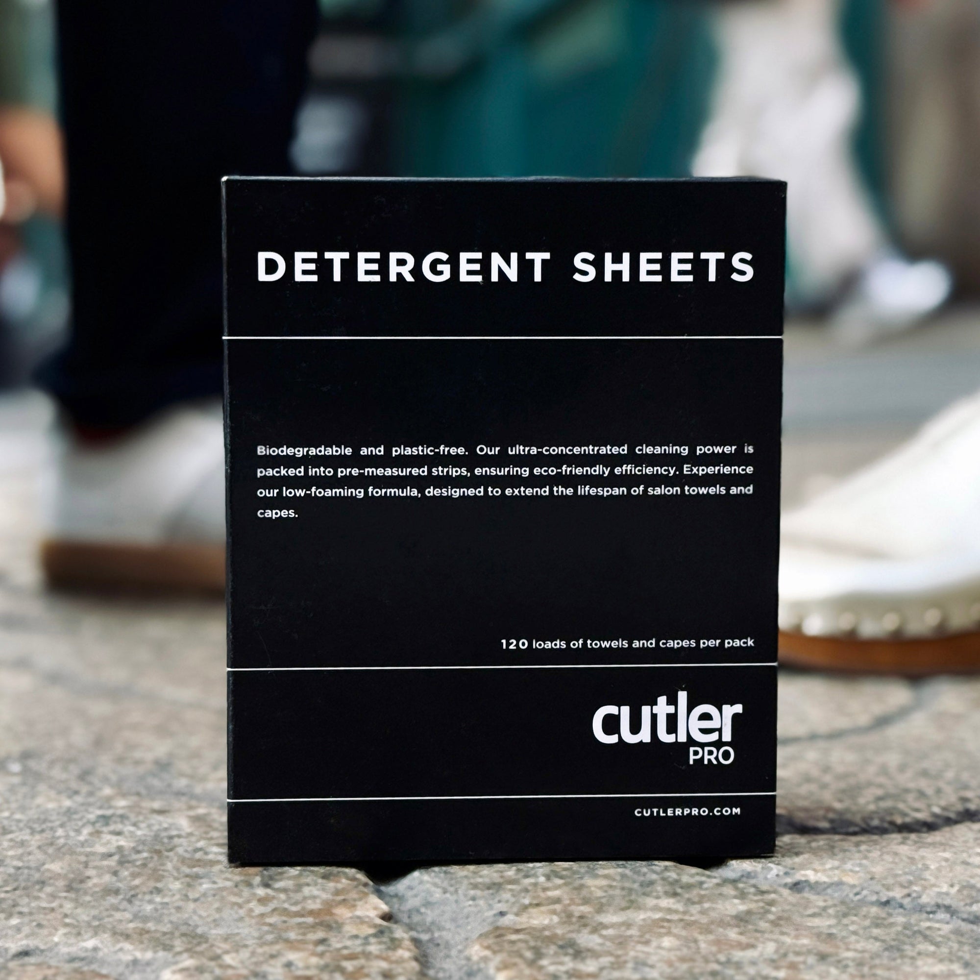 Black detergent sheet box with white writing in front of background of a city sidewalk and people walking.
